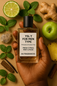 A dark-skinned hand holding a rectangular glass perfume bottle labeled “YSL Y for Men Type – Fresh Citrus, Mint & Musk” by HA Fragrances, surrounded by bergamot, ginger root, mint leaves, green apple, pineapple slice, ambergris, vanilla pod, and cedarwood pieces on a warm beige background